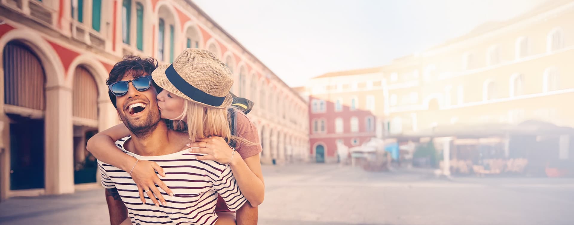 Couple souriant dans une place ensoleillée bordée de bâtiments colorés.